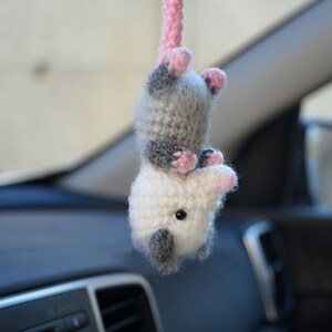 May include: A crocheted plush toy shaped like an opossum hanging from a rearview mirror in a car. The opossum is white and gray with pink ears and a pink nose.
