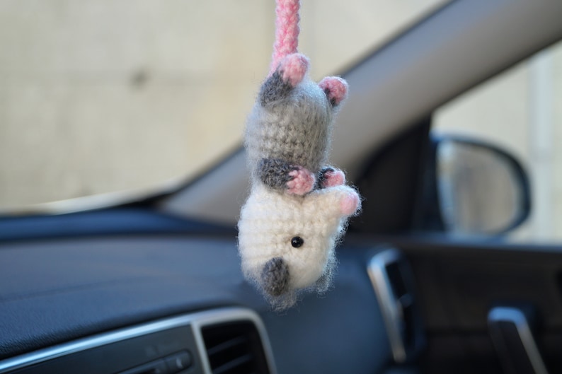May include: A crocheted plush toy shaped like an opossum hanging from a rearview mirror in a car. The opossum is white and gray with pink ears and a pink nose.