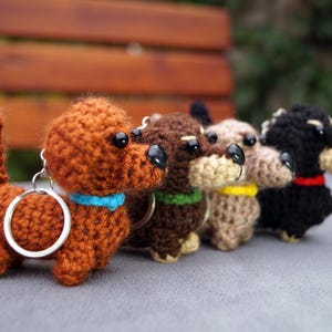 May include: A line of crocheted dachshund keyrings in shades of brown, dark brown, beige, and black. Each dog has a different coloured collar: blue, green, yellow, and red. The keyrings are displayed on a grey surface.