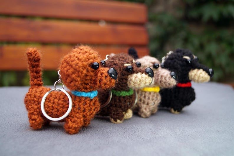 May include: A line of crocheted dachshund keyrings in shades of brown, dark brown, beige, and black. Each dog has a different coloured collar: blue, green, yellow, and red. The keyrings are displayed on a grey surface.