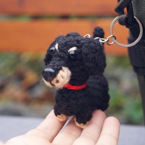 May include: A crocheted black and tan dog keyring with a red collar. The small dog has a black nose and is attached to a silver keyring. The dog is a miniature dachshund.