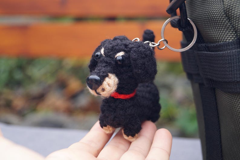May include: A crocheted black and tan dog keyring with a red collar. The small dog has a black nose and is attached to a silver keyring. The dog is a miniature dachshund.