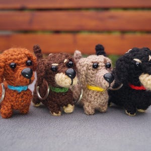 May include: Four crocheted dachshund dog keyrings in shades of brown, dark brown, beige, and black. Each dog has a black nose, small eyes, and a collar in blue, green, yellow, and red. The keyrings have silver hoops.