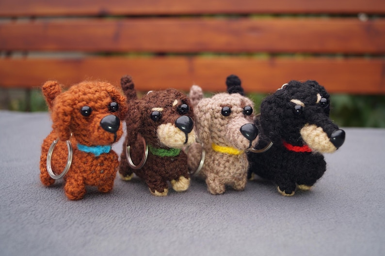May include: Four crocheted dachshund dog keyrings in shades of brown, dark brown, beige, and black. Each dog has a black nose, small eyes, and a collar in blue, green, yellow, and red. The keyrings have silver hoops.