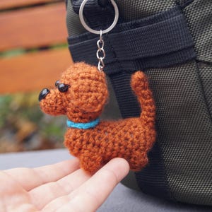 May include: A handmade crocheted dog keyring in shades of brown, with black eyes and a blue collar. The dog charm is attached to a silver keyring. The keyring is shown next to a green bag.