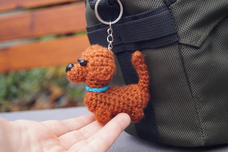 May include: A handmade crocheted dog keyring in shades of brown, with black eyes and a blue collar. The dog charm is attached to a silver keyring. The keyring is shown next to a green bag.