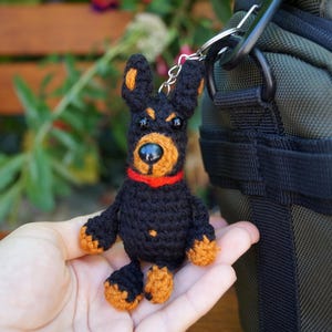 May include: A handmade crochet Doberman Pinscher dog keyring. The dog is black with tan markings on its ears, snout, and paws, and a red collar. The keyring is attached to a silver metal ring.