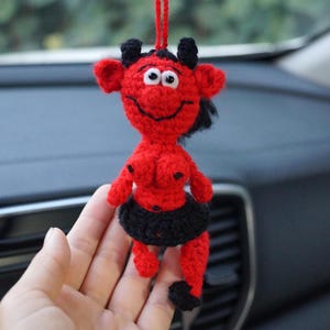 Pode incluir: Um ornamento de diabo em crochê vermelho e preto, apresentando uma pequena figura com chifres pretos, saia preta e ponta da cauda preta. A figura tem olhos brancos com pupilas pretas, um sorriso bordado e um laço de fio vermelho no topo.