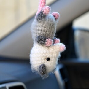 May include: A crocheted plush opossum toy with gray and white fur, pink ears and nose, and black eyes. The toy is hanging from a car's rearview mirror.