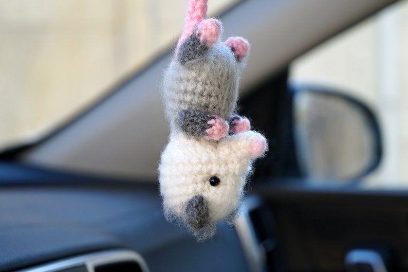 May include: A crocheted plush opossum toy with gray and white fur, pink ears and nose, and black eyes. The toy is hanging from a car's rearview mirror.