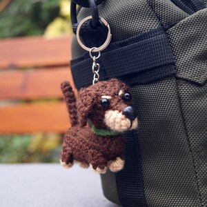 May include: A crocheted dachshund dog keyring in shades of brown, beige, and black, with a green collar. The dog is attached to a silver keyring and chain, hanging next to a dark green bag.