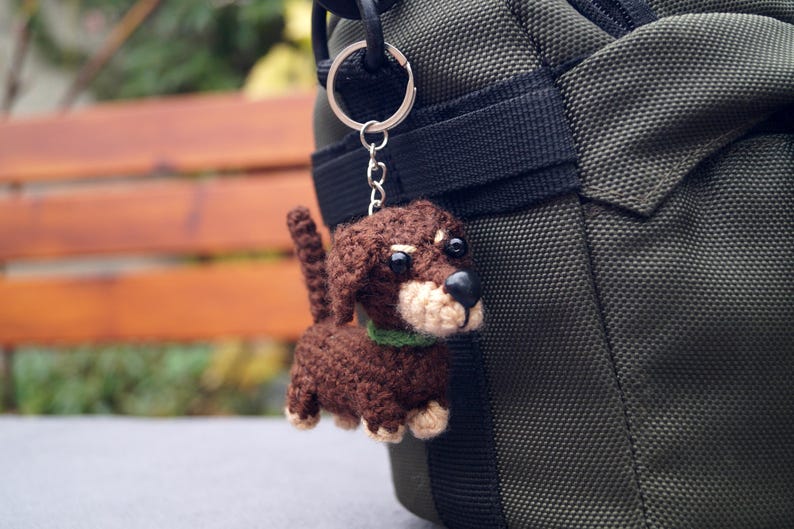 May include: A crocheted dachshund dog keyring in shades of brown, beige, and black, with a green collar. The dog is attached to a silver keyring and chain, hanging next to a dark green bag.