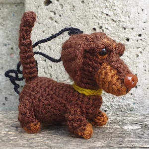 May include: A brown crocheted dachshund toy with a yellow collar. The toy has a black string attached to the top of its head.