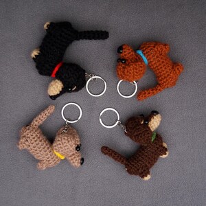 May include: Four handmade crochet dachshund keyrings in black, brown, and tan. Each keyring has a small metal ring. The dachshunds have details like red, yellow, and blue collars.