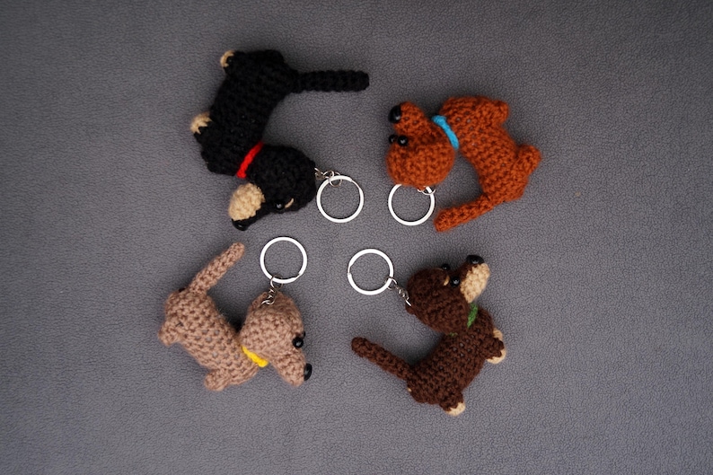 May include: Four handmade crochet dachshund keyrings in black, brown, and tan. Each keyring has a small metal ring. The dachshunds have details like red, yellow, and blue collars.