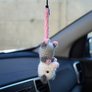 May include: A crocheted gray and pink opossum hanging from a rearview mirror in a car. The opossum has pink ears and a pink nose.