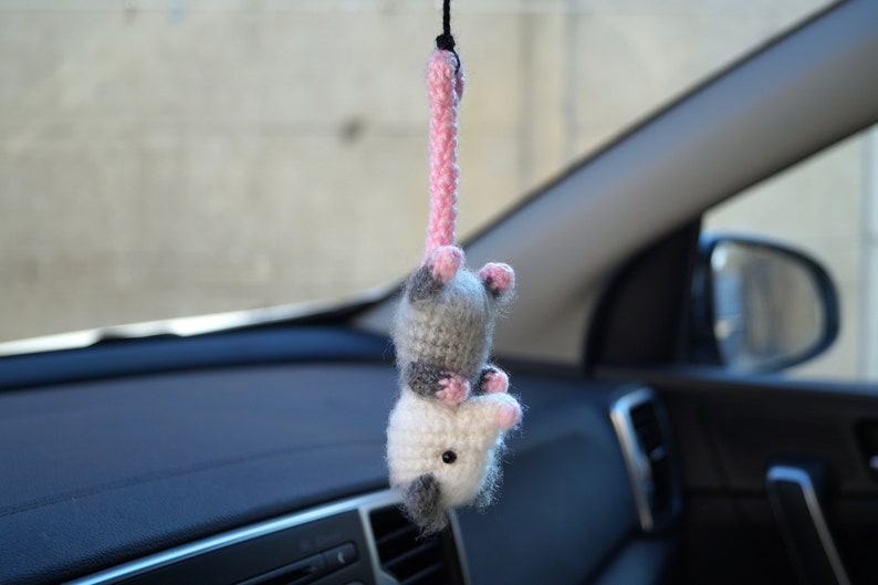 May include: A crocheted gray and pink opossum hanging from a rearview mirror in a car. The opossum has pink ears and a pink nose.