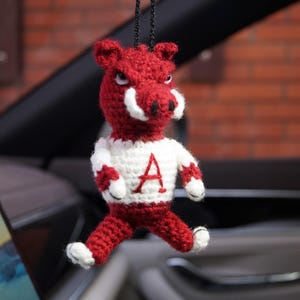 May include: A crocheted hanging ornament shaped like a maroon and white boar mascot. The figure features a maroon head with white tusks, a white torso with a red letter A on the chest, and maroon limbs with white hands and feet, suspended by a black cord.