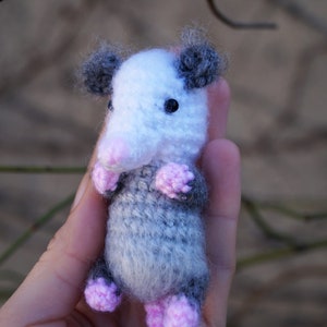 May include: A small, crocheted opossum plush toy with gray and white fur, pink ears, and black eyes. The toy is being held in a person's hand.