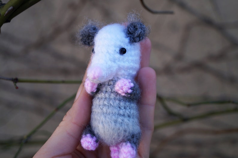 May include: A small, crocheted opossum plush toy with gray and white fur, pink ears, and black eyes. The toy is being held in a person's hand.