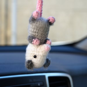 May include: A gray and white crocheted opossum hanging from a rearview mirror in a car. The opossum has pink ears and a pink nose.