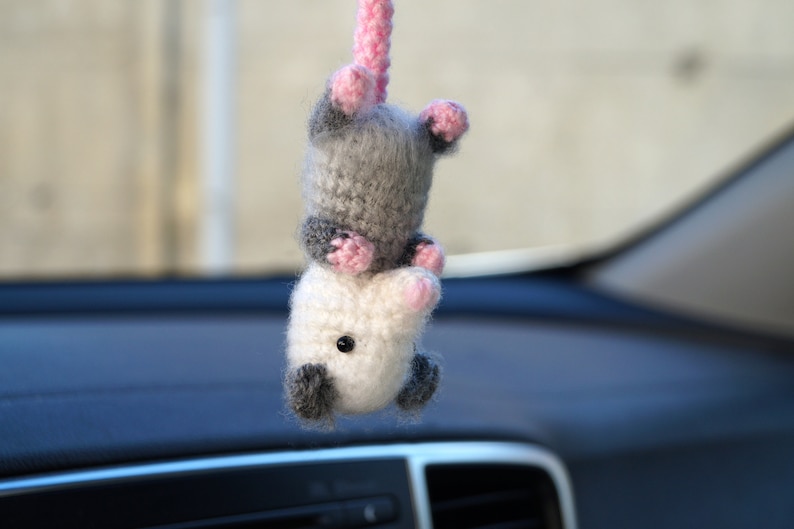 May include: A gray and white crocheted opossum hanging from a rearview mirror in a car. The opossum has pink ears and a pink nose.