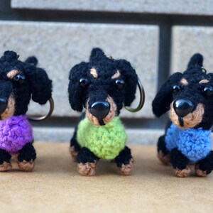 Może przedstawiać: Trzy breloczki z dzianych ps&oacute;w rasy dachshund. Dachshunds są czarne z brązowymi nosami i mają r&oacute;żne kolory swetr&oacute;w: fioletowy, zielony i niebieski.