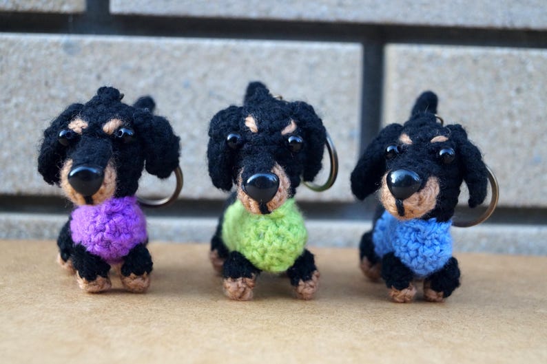 Może przedstawiać: Trzy breloczki z dzianych ps&oacute;w rasy dachshund. Dachshunds są czarne z brązowymi nosami i mają r&oacute;żne kolory swetr&oacute;w: fioletowy, zielony i niebieski.