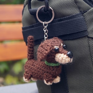 May include: A crocheted dachshund dog keyring in shades of brown, beige, and black, with a green collar. The dog is attached to a silver keyring and chain, hanging against a green fabric background.