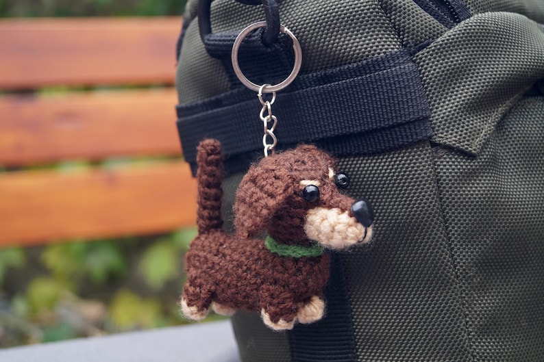 May include: A crocheted dachshund dog keyring in shades of brown, beige, and black, with a green collar. The dog is attached to a silver keyring and chain, hanging against a green fabric background.