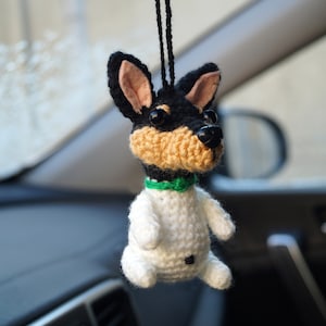 May include: A crocheted dog toy hanging from a rearview mirror in a car. The dog is white with black and brown accents and a green bow tie.