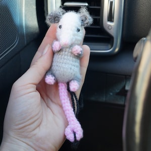 May include: A small gray and white crocheted opossum with pink ears and a pink tail. The opossum is hanging from a black string.