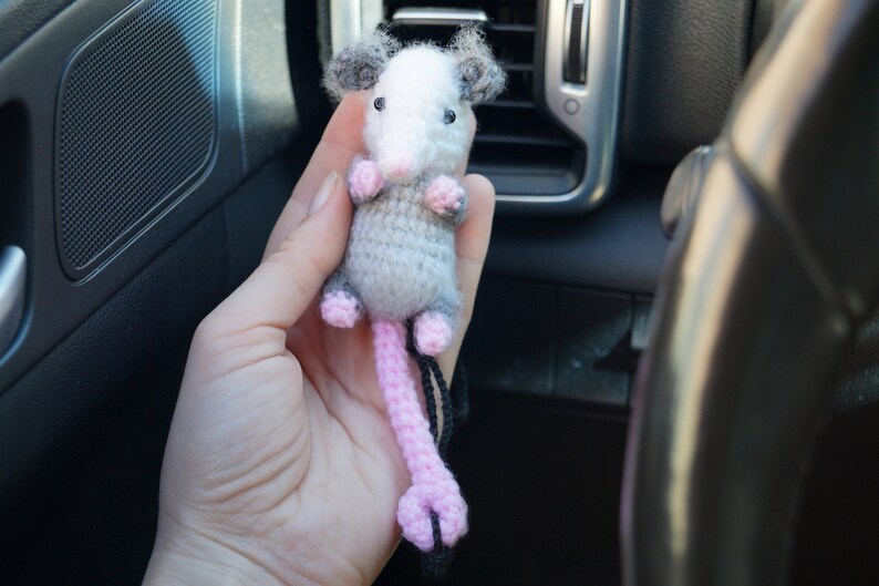 May include: A small gray and white crocheted opossum with pink ears and a pink tail. The opossum is hanging from a black string.