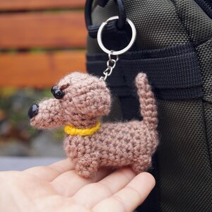 May include: A crocheted dachshund dog keyring in shades of brown, with black eyes and a yellow collar. The dog is attached to a silver keyring. The keyring is held in front of a green bag.