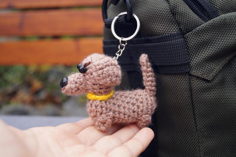 May include: A crocheted dachshund dog keyring in shades of brown, with black eyes and a yellow collar. The dog is attached to a silver keyring. The keyring is held in front of a green bag.