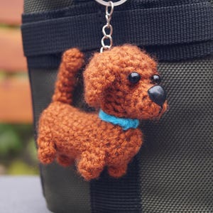 May include: A crocheted dog keyring in shades of brown, with a blue collar and black eyes and nose. The handmade dog is attached to a silver keyring and hangs against a dark green background.