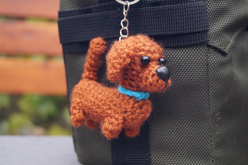 May include: A crocheted dog keyring in shades of brown, with a blue collar and black eyes and nose. The handmade dog is attached to a silver keyring and hangs against a dark green background.