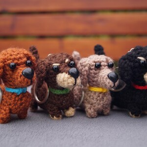 May include: Four crocheted dachshund keyrings in shades of brown and black. Each dog has a black nose and a coloured collar in blue, green, yellow, and red. The keyrings are displayed on a grey surface.