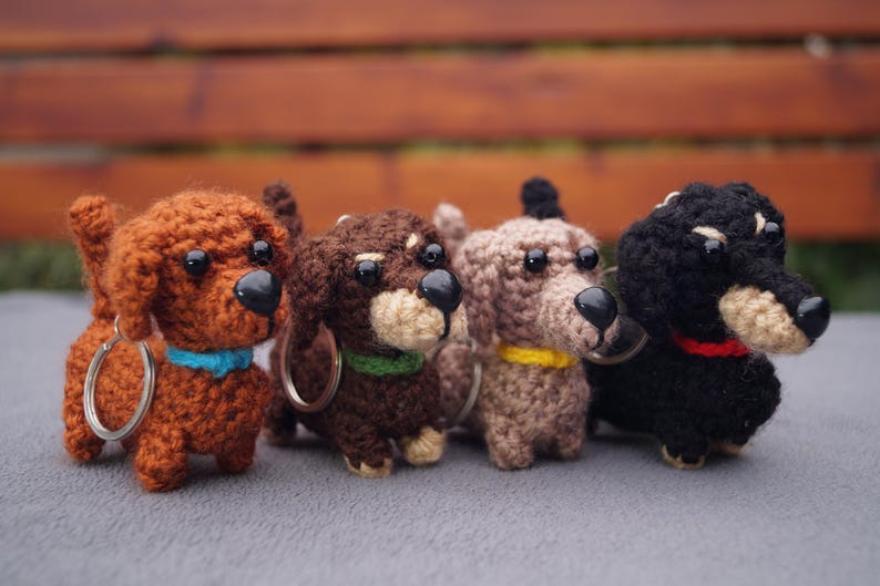 May include: Four crocheted dachshund keyrings in shades of brown and black. Each dog has a black nose and a coloured collar in blue, green, yellow, and red. The keyrings are displayed on a grey surface.