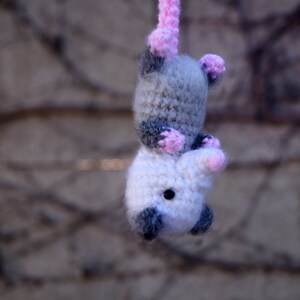 May include: A crocheted gray and white opossum with pink ears and nose. The opossum is hanging from a string.