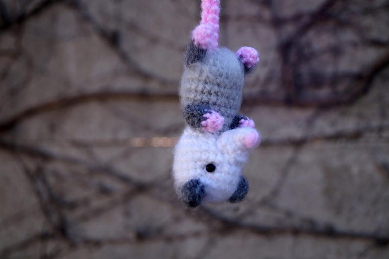 May include: A crocheted gray and white opossum with pink ears and nose. The opossum is hanging from a string.