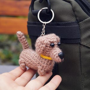 May include: A crocheted dachshund dog keyring in shades of brown, with a yellow collar and black eyes and nose. The dog is attached to a silver keyring. The background includes a green bag and a wooden bench.