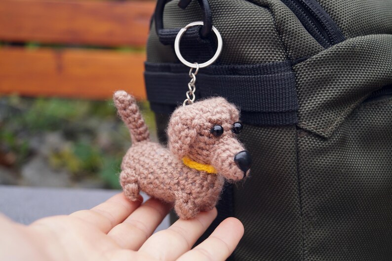 May include: A crocheted dachshund dog keyring in shades of brown, with a yellow collar and black eyes and nose. The dog is attached to a silver keyring. The background includes a green bag and a wooden bench.