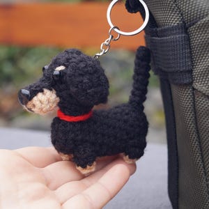May include: A crocheted dachshund keyring in black with tan detailing and a red collar. The small dog has a silver keyring attached. The keyring is shown next to a green bag.