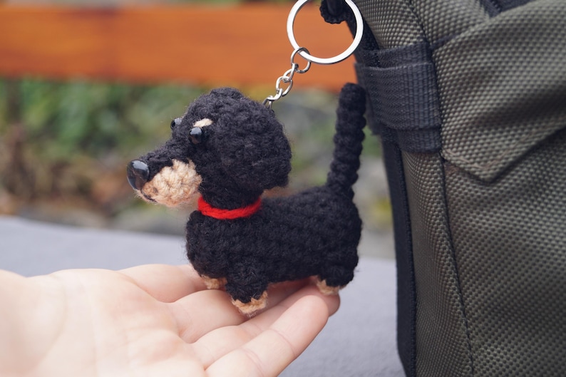 May include: A crocheted dachshund keyring in black with tan detailing and a red collar. The small dog has a silver keyring attached. The keyring is shown next to a green bag.