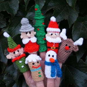 May include: A set of seven crocheted finger puppets featuring holiday characters including a green elf, a grey-haired figure, a green Christmas tree with red dots, Santa Claus, a brown reindeer, a gingerbread person, and a white snowman with a blue scarf.