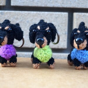 Może przedstawiać: Trzy breloczki z dzianych ps&oacute;w rasy dachshund w swetrach w r&oacute;żnych kolorach. Dachshund po lewej stronie ma na sobie fioletowy sweter, dachshund pośrodku ma na sobie zielony sweter, a dachshund po prawej stronie ma na sobie niebieski sweter.