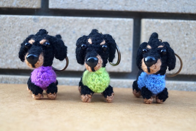 Może przedstawiać: Trzy breloczki z dzianych ps&oacute;w rasy dachshund w swetrach w r&oacute;żnych kolorach. Dachshund po lewej stronie ma na sobie fioletowy sweter, dachshund pośrodku ma na sobie zielony sweter, a dachshund po prawej stronie ma na sobie niebieski sweter.