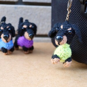 Może przedstawiać: Trzy breloczki z dzianych ps&oacute;w rasy dachshund. Jeden ma na sobie niebieski sweter, drugi fioletowy, a trzeci zielony.