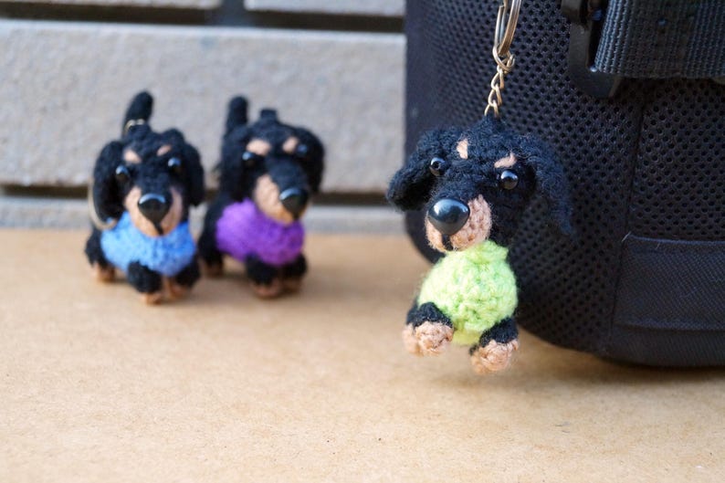 Może przedstawiać: Trzy breloczki z dzianych ps&oacute;w rasy dachshund. Jeden ma na sobie niebieski sweter, drugi fioletowy, a trzeci zielony.